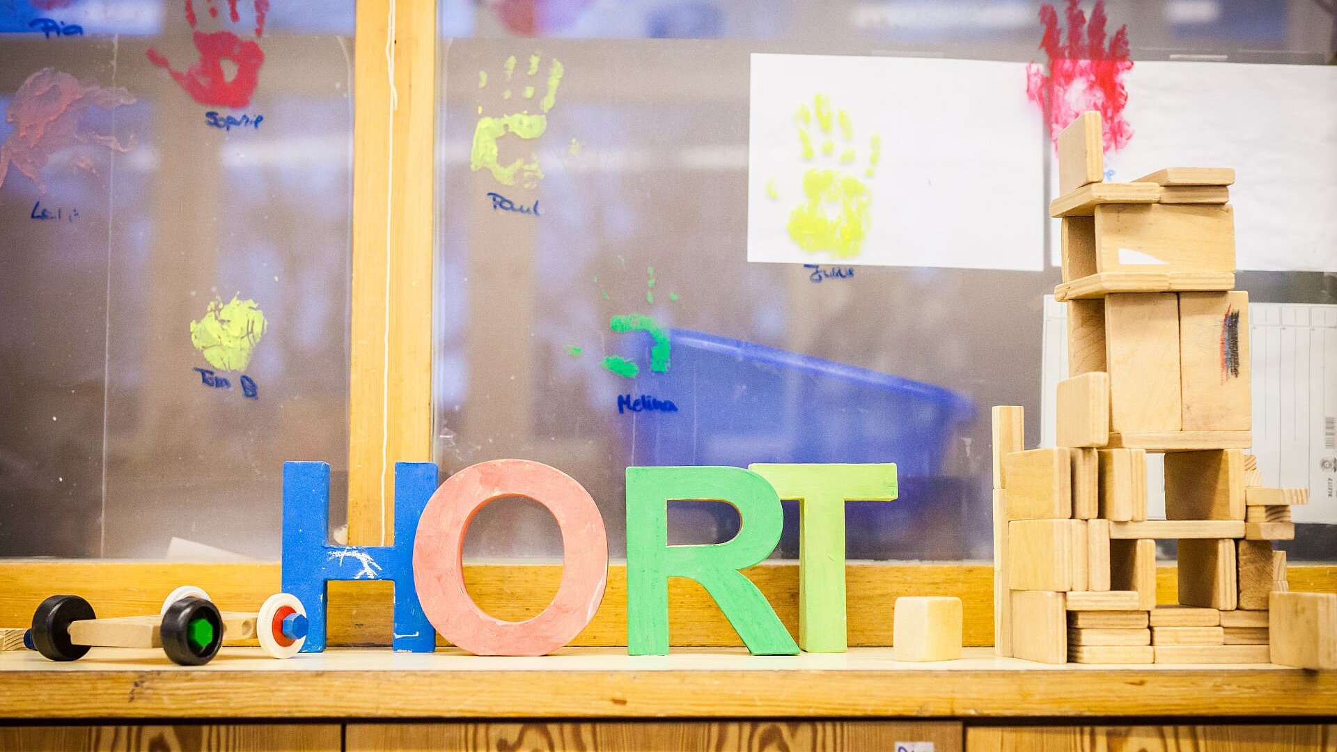 Schriftzug HORT in bunten Buchstaben vor einem Fenster mit bunten Handabdrücken, daneben Holzspielzeug.