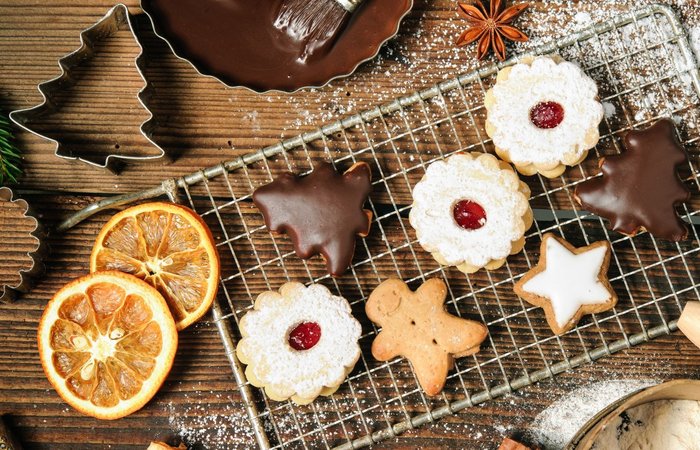 Verschiedene weihnachtliche Kekse, darunter mit Schokolade überzogene, mit Puderzucker bestreute und sternförmige, liegen auf einem Gitterrost. Drumherum liegen getrocknete Orangenscheiben, Ausstechformen und Tannenzweige auf einer Holzoberfläche.