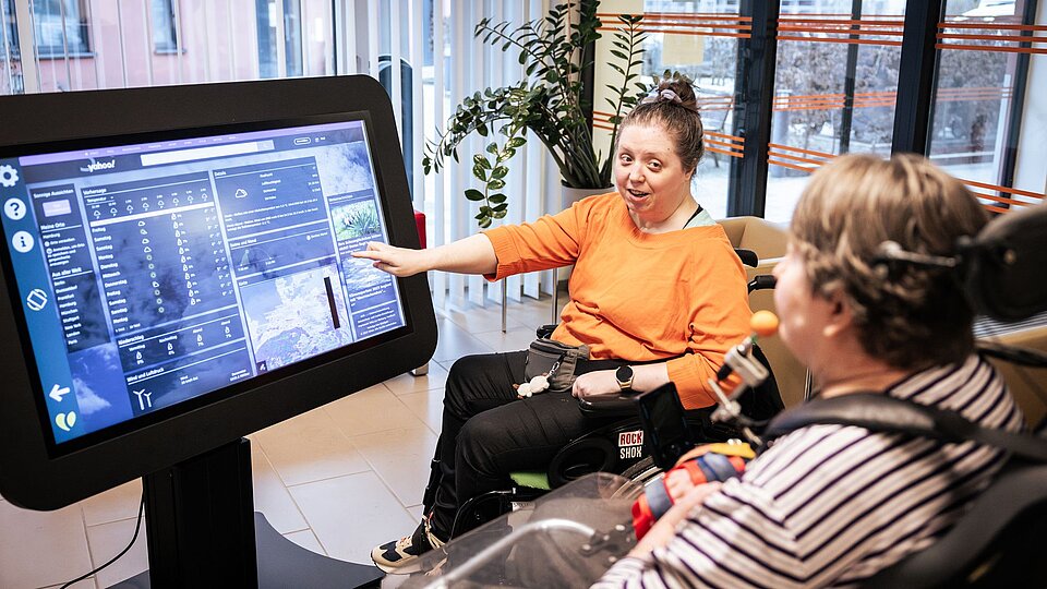 Zwei Frauen im Rollstuh sitzen um einen großen Bildschirm, der eine Internetseite mit dem Wetter zeigt. Eine zeigt auf den Bildschirm. 