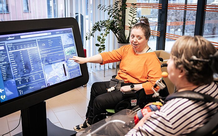 Zwei Frauen im Rollstuh sitzen um einen großen Bildschirm, der eine Internetseite mit dem Wetter zeigt. Eine zeigt auf den Bildschirm.