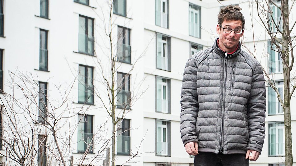 Ein Spieler der Boule Gruppe Baaken Bande in einer warmen Jacke. Im Hintergrund ein Wohnhaus in der HafenCity.