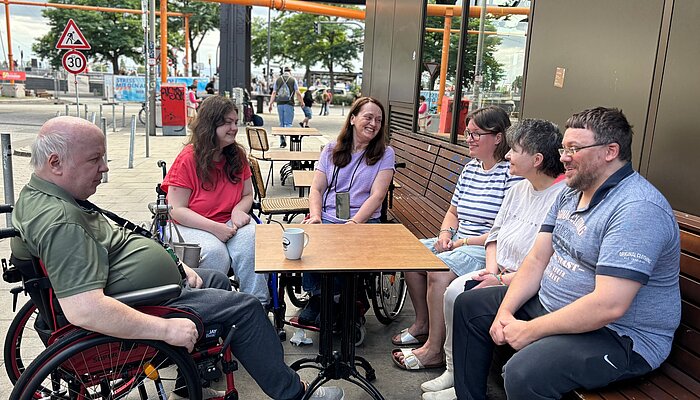 Sechs Teilnehmende des Hafentreffs sitzen gemeinsam an einem Tisch vor der Schanzenbäckerei nahe den Landungsbrücken. Zwei Personen nutzen einen Rollstuhl, die Gruppe ist im Gespräch und lacht miteinander.