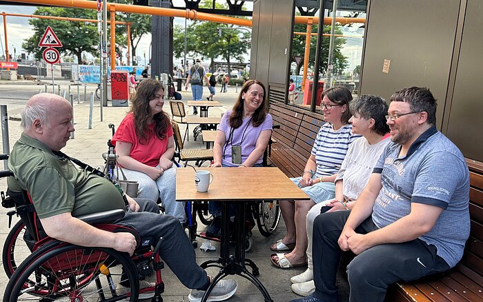 Sechs Teilnehmende des Hafentreffs sitzen gemeinsam an einem Tisch vor der Schanzenbäckerei nahe den Landungsbrücken. Zwei Personen nutzen einen Rollstuhl, die Gruppe ist im Gespräch und lacht miteinander.