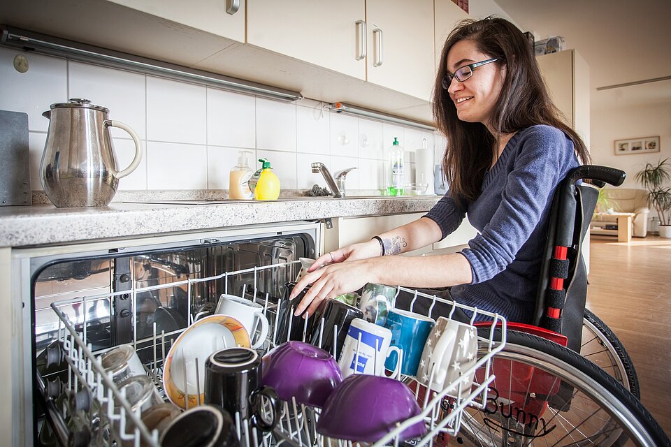 Eine Person sitzt in einem Rollstuhl vor einer geöffneten Spülmaschine und räumt Geschirr in den unteren Korb ein. Die Szene spielt in einer barrierefrei gestalteten Küche mit unterfahrbarer Arbeitsfläche.