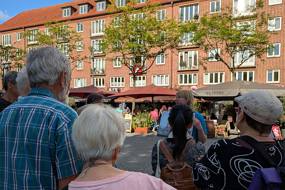 Eine Gruppe Menschen steht im Halbkreis um die Stadtführerin, die etwas in der Hand hält.