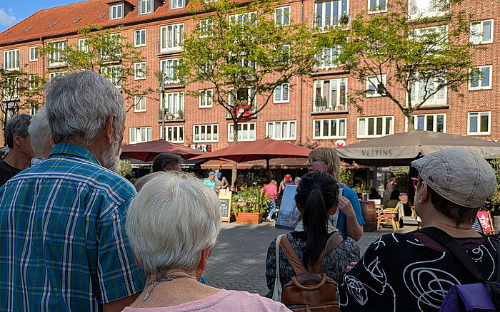 Eine Gruppe Menschen steht im Halbkreis um die Stadtführerin, die etwas in der Hand hält.