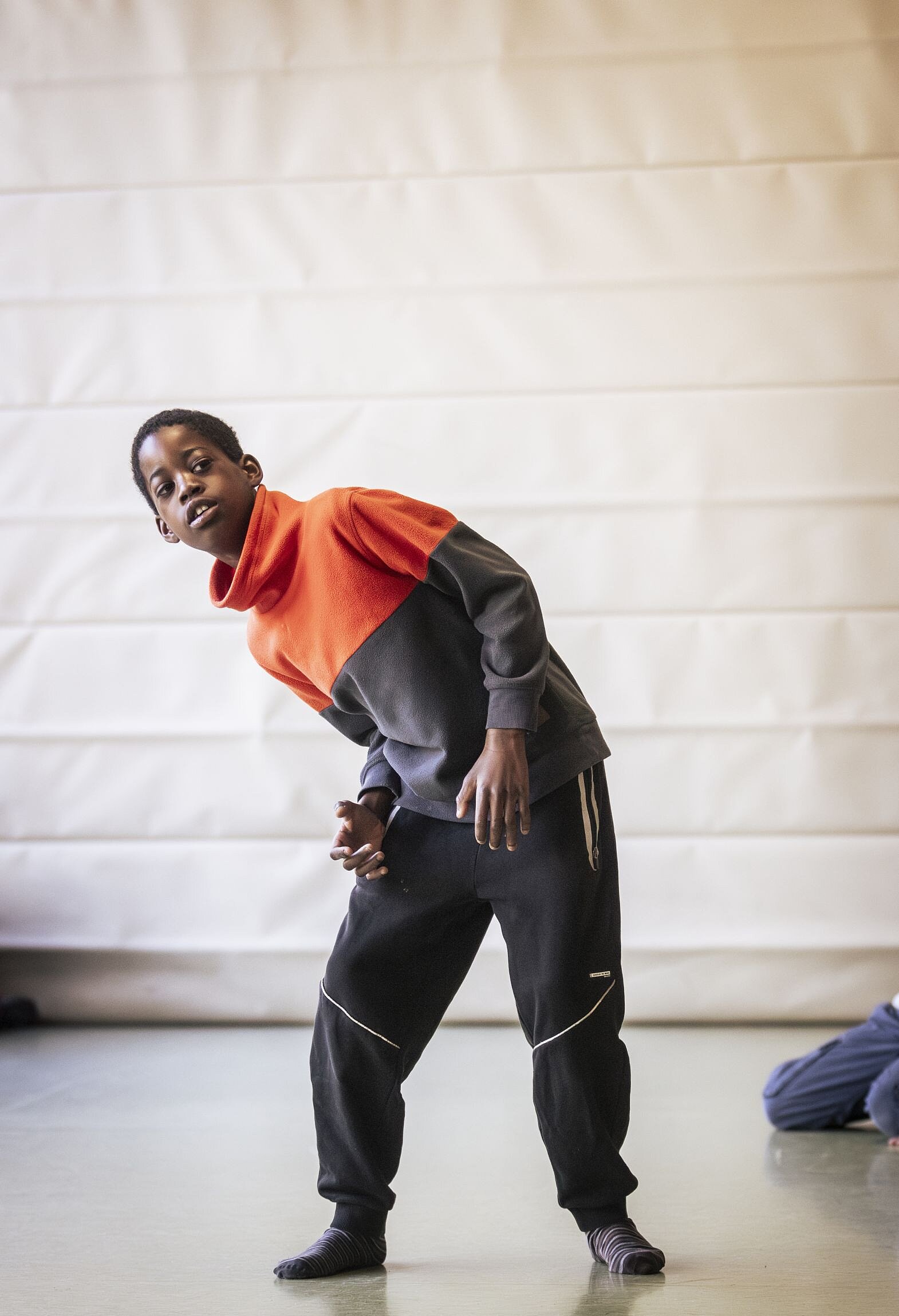 Ein Junge in einem zweifarbigen Trainingsanzug beugt sich tänzerisch zur Seite. Er schaut direkt in die Kamera und wirkt konzentriert und ausdrucksstark. Der Hintergrund ist eine weiche Hallenwand.
