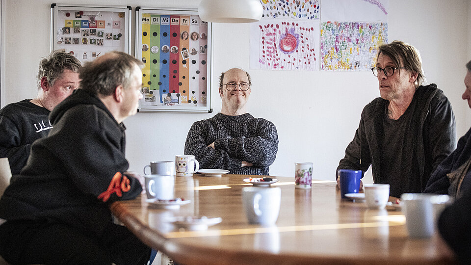 Fünf Erwachsene sitzen um einen runden Holztisch in einem hellen Raum. Sie trinken Kaffee aus bunten Tassen und unterhalten sich. Im Hintergrund sind an der Wand selbstgemalte Bilder und ein bunter Wochenplan zu sehen.