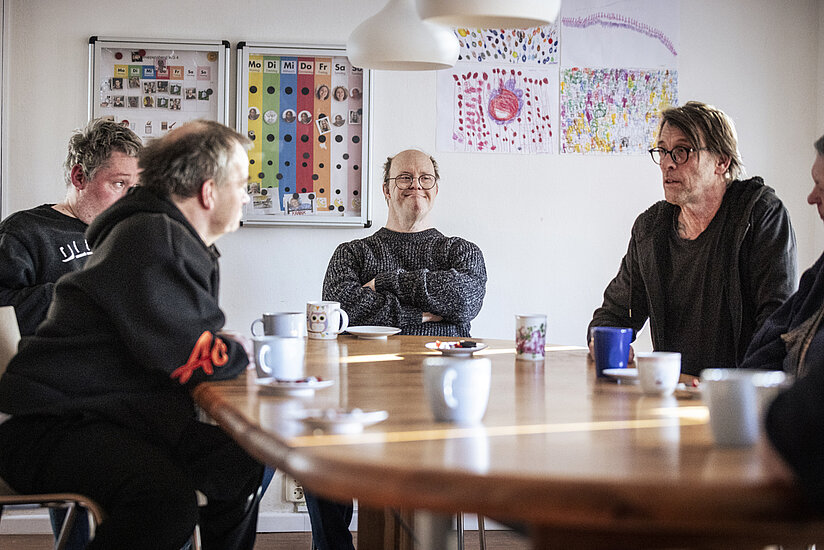 Fünf Erwachsene sitzen um einen runden Holztisch in einem hellen Raum. Sie trinken Kaffee aus bunten Tassen und unterhalten sich. Im Hintergrund sind an der Wand selbstgemalte Bilder und ein bunter Wochenplan zu sehen.