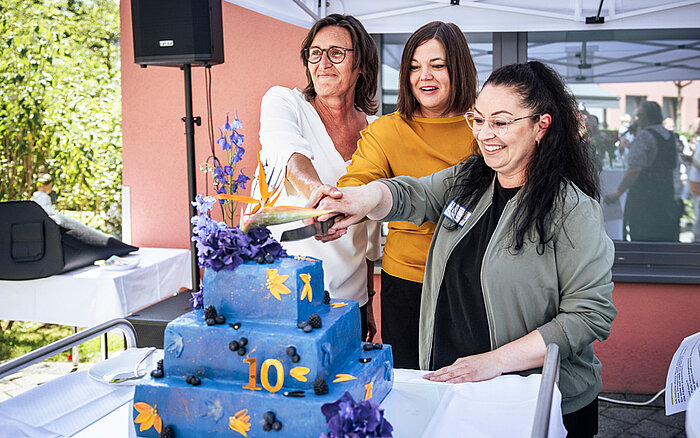 Drei Frauen (darunter Katharina Fegebank, Hamburgs Zweite Bürgermeisterin und Ina Terragnolo, die Leiterin des Haus Beerboom sowie eine weitere Mitarbeitende des Haus Beerboom) stehen gemeinsam an einem Tisch unter einem Pavillon und schneiden eine große, mehrstöckige Torte an. Die Torte ist blau mit bunten Zahlen und Symbolen dekoriert, obenauf blauer und lilafarbener Blumenschmuck. Im Hintergrund sind ein Gebäude und etwas Grün zu erkennen.