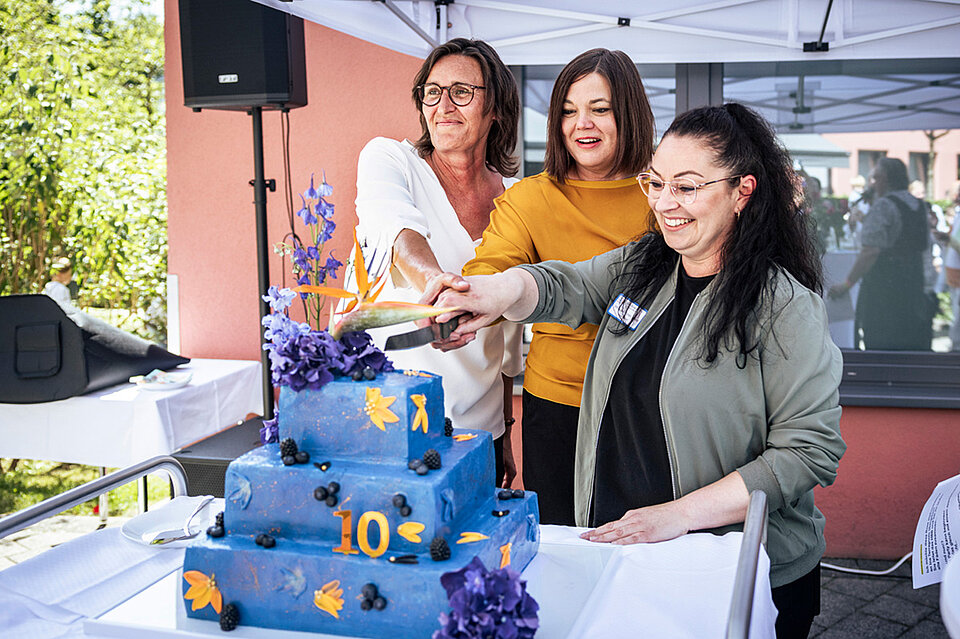 Drei Frauen (darunter Katharina Fegebank, Hamburgs Zweite Bürgermeisterin und Ina Terragnolo, die Leiterin des Haus Beerboom sowie eine weitere Mitarbeitende des Haus Beerboom) stehen gemeinsam an einem Tisch unter einem Pavillon und schneiden eine große, mehrstöckige Torte an. Die Torte ist blau mit bunten Zahlen und Symbolen dekoriert, obenauf blauer und lilafarbener Blumenschmuck. Im Hintergrund sind ein Gebäude und etwas Grün zu erkennen.