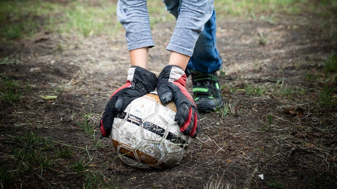 Ein Fußball liegt im Dreck und wird von einem Kind mit Torwarthandschuhen aufgehoben