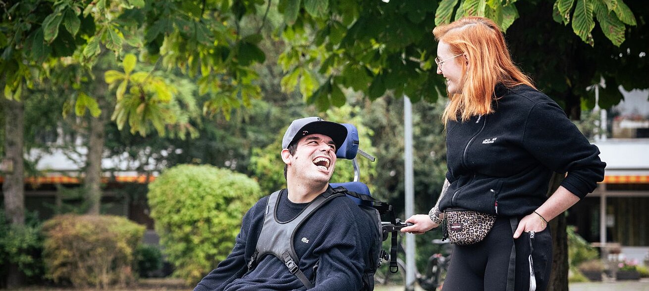 Eine Person im Rollstuhl und eine junge Assistenzkraft unterhalten sich lachend im Grünen. Die Assistenzkraft steht neben dem Rollstuhl und hält die Hand locker am Griff.