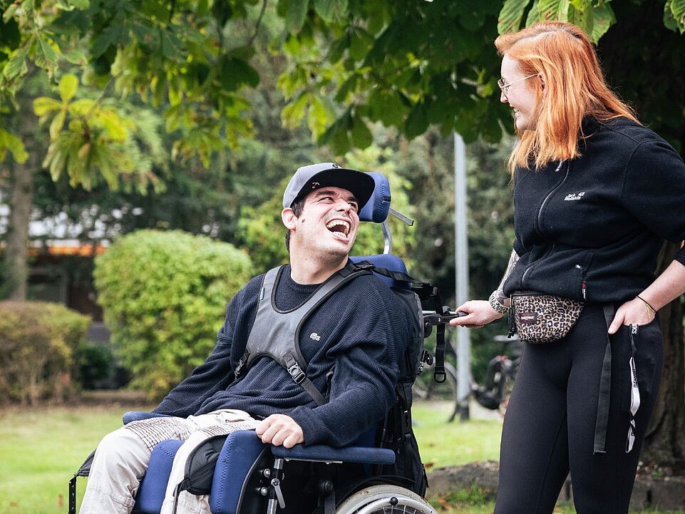 Eine Person im Rollstuhl und eine junge Assistenzkraft unterhalten sich lachend im Grünen. Die Assistenzkraft steht neben dem Rollstuhl und hält die Hand locker am Griff.