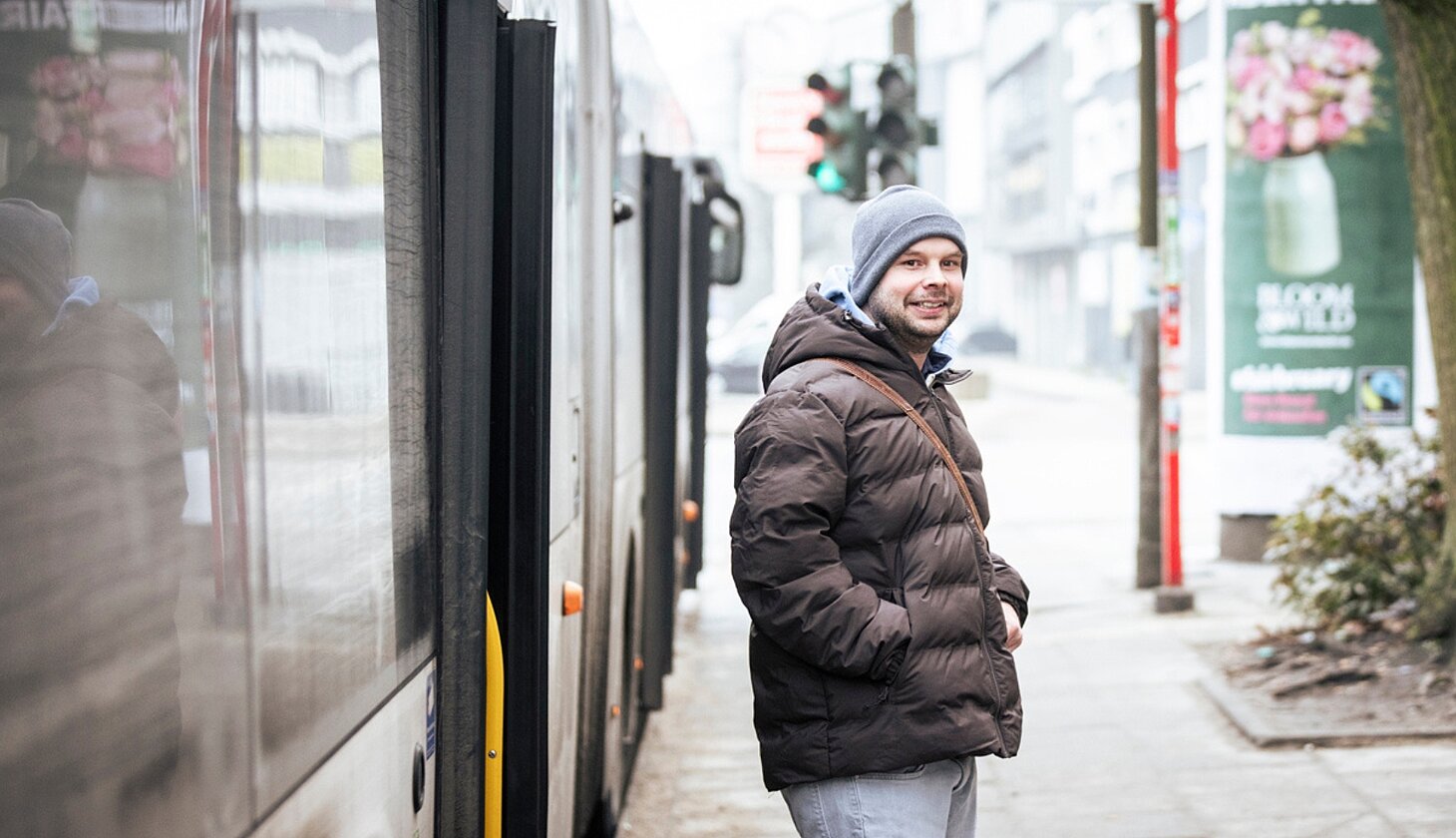 Niklas Luy steht an einer Bushaltestelle neben einem Bus und lächelt in die Kamera. Er trägt eine Winterjacke und eine Mütze.