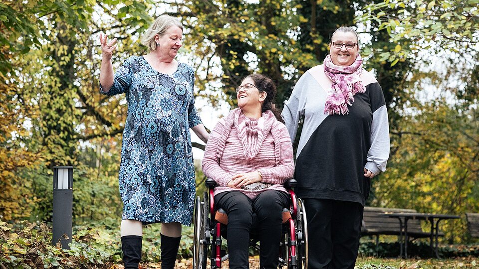 Drei Frauen im Grünen, die Frau in der Mitte sitzt im Rollstuhl.