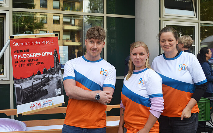 Drei Personen in orange-blauen Shirts stehen vor einem Gebäude neben einem Plakat zur Pflegereform.