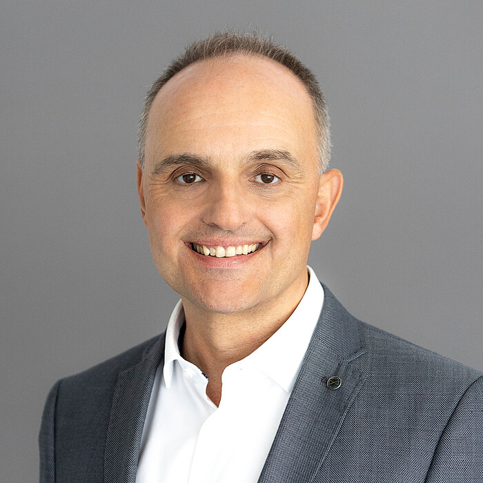 Michael Wild, ein Mann mit kurzem, grauem Haar und Brille, lächelt freundlich in die Kamera. Er trägt ein gemustertes Sakko und ein weißes Hemd. Der Hintergrund ist dunkelgrau gestaltet.