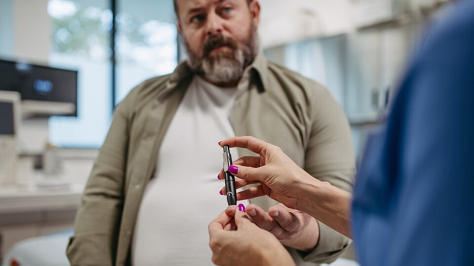 Mittelalter Mann mit Übergewicht, bei dem eine medizinische Fachkraft Blut aus dem Finger für einen Blutzucker-Test abnimmt.
