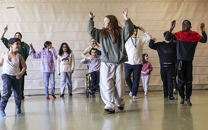 Eine Gruppe von Kindern und Jugendlichen, darunter auch Teilnehmende im Rollstuhl, tanzt gemeinsam in einer großen Turnhalle. Die Bewegungen folgen einem choreografierten Ablauf, die Arme sind erhoben. Die zentrale Tänzerin führt die Gruppe an.