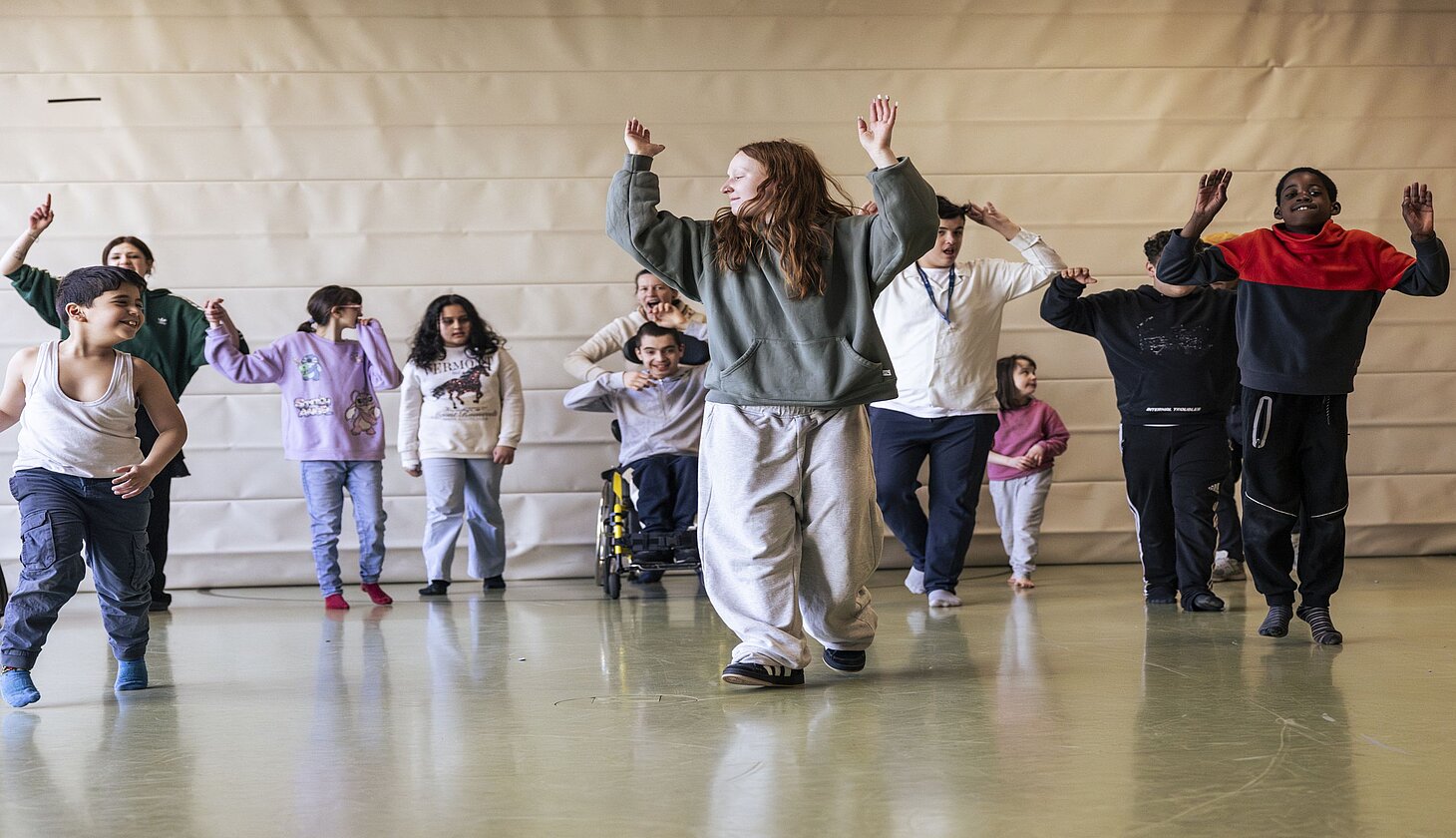 Eine Gruppe von Kindern und Jugendlichen, darunter auch Teilnehmende im Rollstuhl, tanzt gemeinsam in einer großen Turnhalle. Die Bewegungen folgen einem choreografierten Ablauf, die Arme sind erhoben. Die zentrale Tänzerin führt die Gruppe an. 