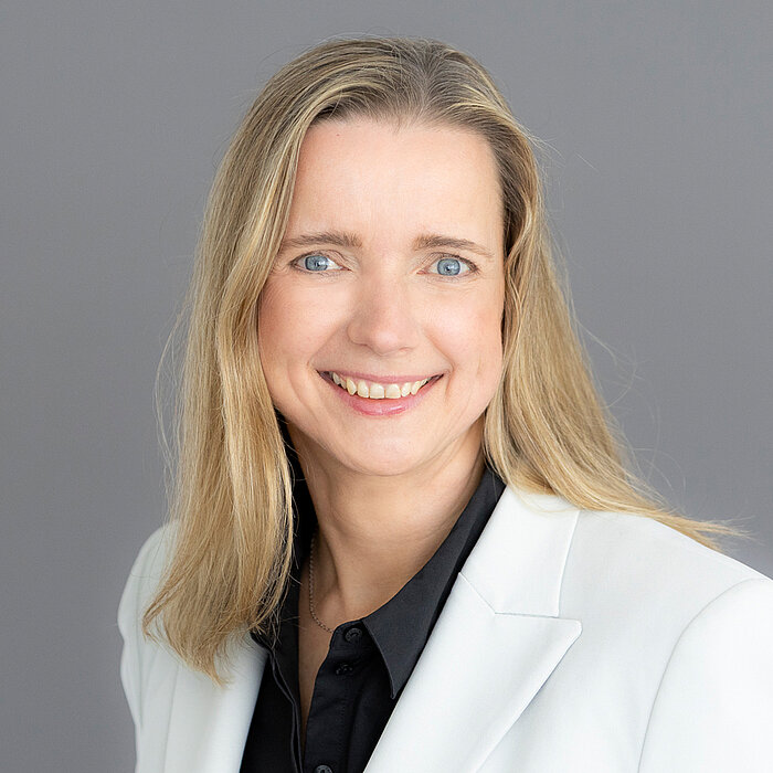 Karin Conradt, eine Frau mit langem, glatten, blonden Haar, lächelt freundlich in die Kamera. Sie trägt ein weißes Hemd unter einem schwarzen Blazer. Der Hintergrund ist neutral grau gestaltet.