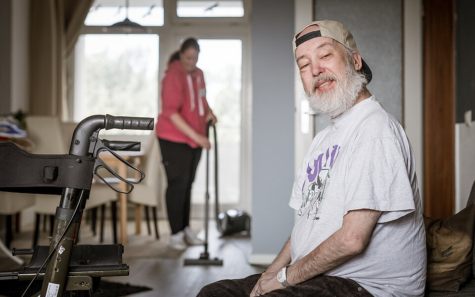 Ein älterer Mann mit grauem Bart, weißem T-Shirt und rückwärts getragener Kappe sitzt in einem Wohnzimmer und schaut freundlich in die Kamera. Im Hintergrund saugt eine Assistenzkraft den Boden. Ein Rollator steht neben dem Mann.