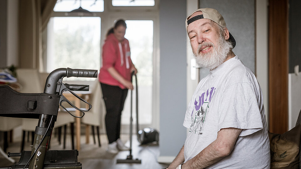 Ein älterer Mann mit grauem Bart, weißem T-Shirt und rückwärts getragener Kappe sitzt in einem Wohnzimmer und schaut freundlich in die Kamera. Im Hintergrund saugt eine Assistenzkraft den Boden. Ein Rollator steht neben dem Mann.
