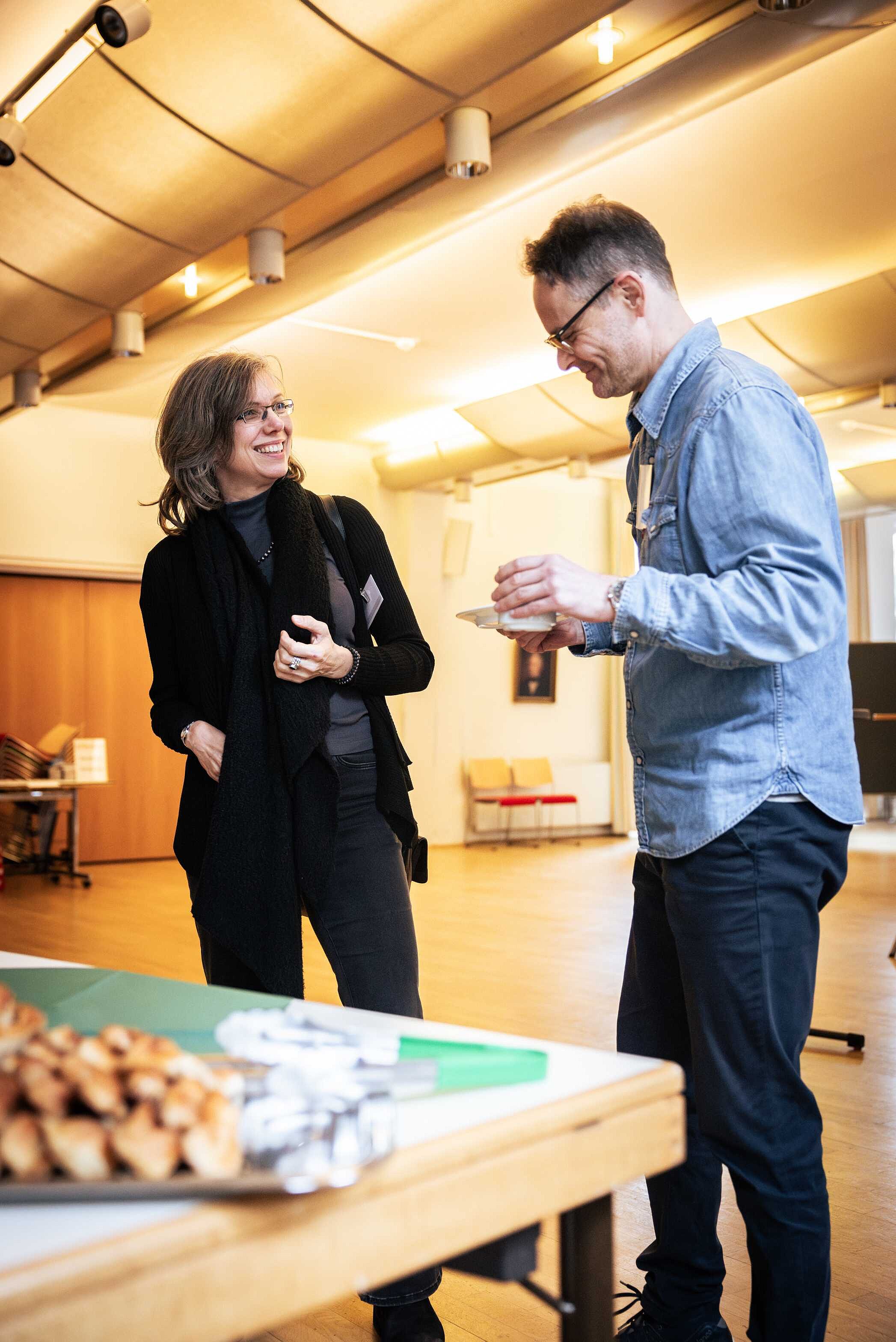 Zwei Personen unterhalten sich entspannt während einer Kaffeepause in einem großen Veranstaltungsraum mit Stühlen im Hintergrund, beide lachen. Links steht eine Frau mit grauen Haaren, Brille und schwarzer Kleidung. Rechts steht ein Mann mit Kaffeetasse in der Hand, grauem Haar, Brille und dunkelblauer Kleidung. Auf dem Tisch im Vordergrund steht Gebäck bereit.
