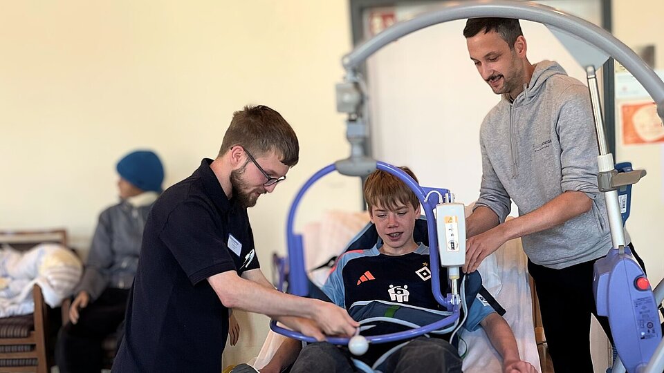 Zwei junge Männer demonstrieren einem Jungen den Einsatz eines mobilen Lifts zur Unterstützung bei der Pflege. Der Junge sitzt in einer Trageschlaufe, die am Lift befestigt ist. Im Hintergrund sind unscharf weitere Personen zu sehen.