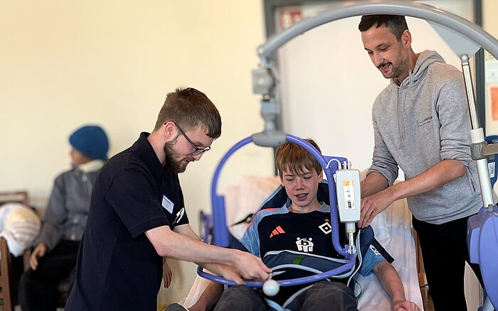 Zwei junge Männer demonstrieren einem Jungen den Einsatz eines mobilen Lifts zur Unterstützung bei der Pflege. Der Junge sitzt in einer Trageschlaufe, die am Lift befestigt ist. Im Hintergrund sind unscharf weitere Personen zu sehen.