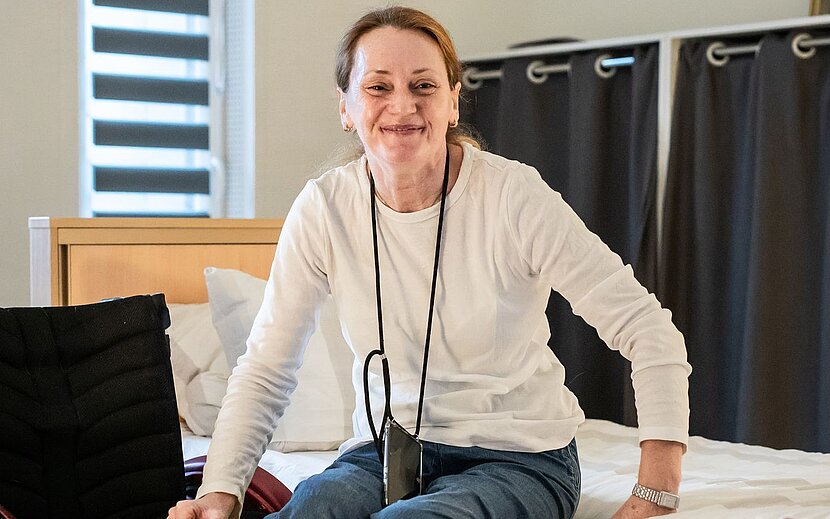 Eine Frau sitzt lächelnd auf einem Bett in einem hellen, modern eingerichteten Zimmer. Sie trägt ein weißes Oberteil und ein Handy mit Band um den Hals.