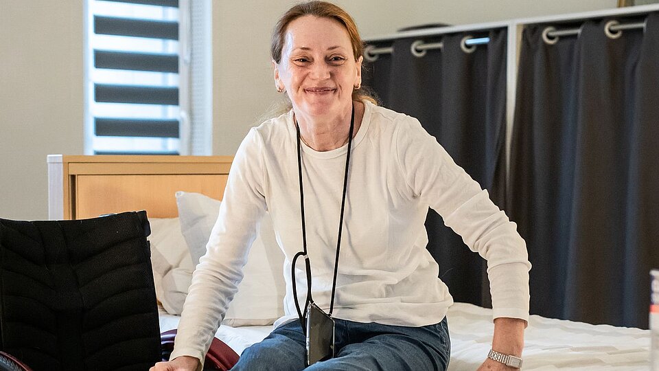 Eine Frau sitzt lächelnd auf einem Bett in einem hellen, modern eingerichteten Zimmer. Sie trägt ein weißes Oberteil und ein Handy mit Band um den Hals.