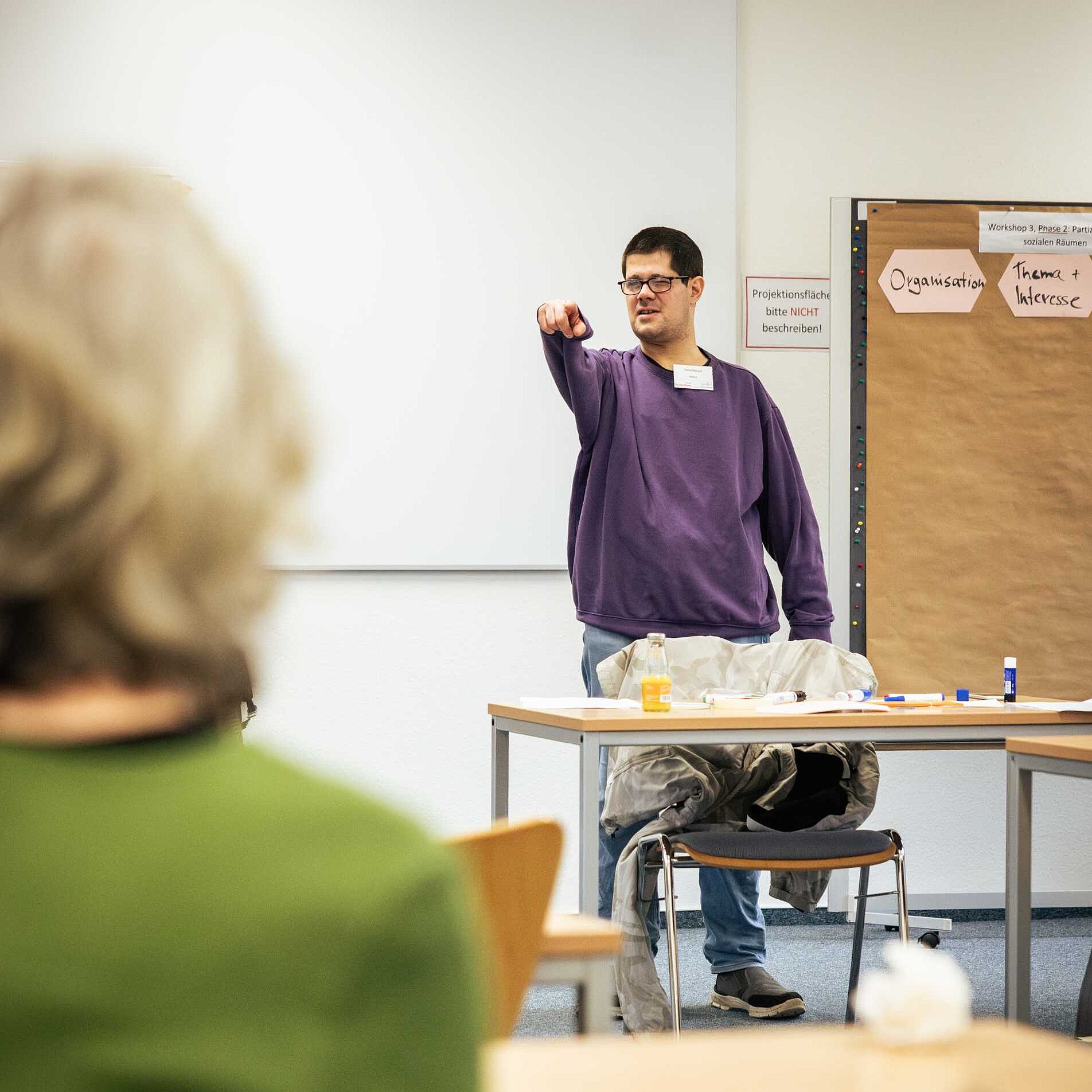 Ein Mann im lila Pullover, kurzem schwarzen Haar, Brille und Namensschild steht an einem Tisch und zeigt gestikulierend in den Raum. Im Vordergrund ist der unscharfe Rücken einer zuhörenden Person zu sehen. Im Hintergrund eine Pinnwand mit der Überschrift „Workshop 3 Phase 2: Partizipation in sozialen Räumen“. Auf den Moderationskarten steht „Organisation“ und „Thema + Interesse“.