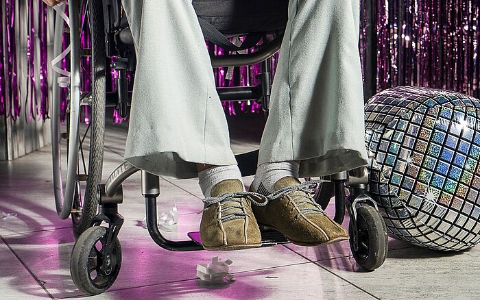 Eine Person sitzt im Rollstuhl auf einer Tanzfläche in Partykleidung. Im Hintergrund ist glitzernde Dekoration und eine Diskokugel zu sehen.