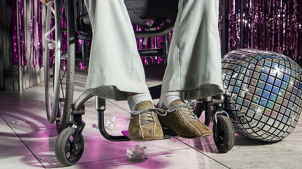 Eine Person sitzt im Rollstuhl auf einer Tanzfläche in Partykleidung. Im Hintergrund ist glitzernde Dekoration und eine Diskokugel zu sehen.