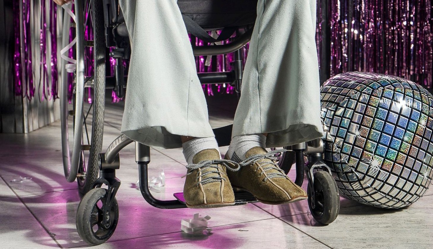 Eine Person sitzt im Rollstuhl auf einer Tanzfläche in Partykleidung. Im Hintergrund ist glitzernde Dekoration und eine Diskokugel zu sehen.