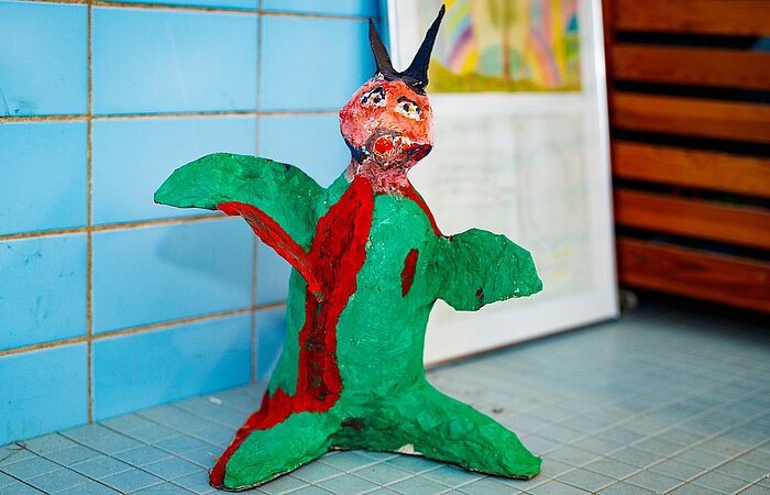 Eine farbenfrohe, handgefertigte Skulptur steht auf einem gefliesten Boden vor einer blau gefliesten Wand mit Kunstwerken im Hintergrund.