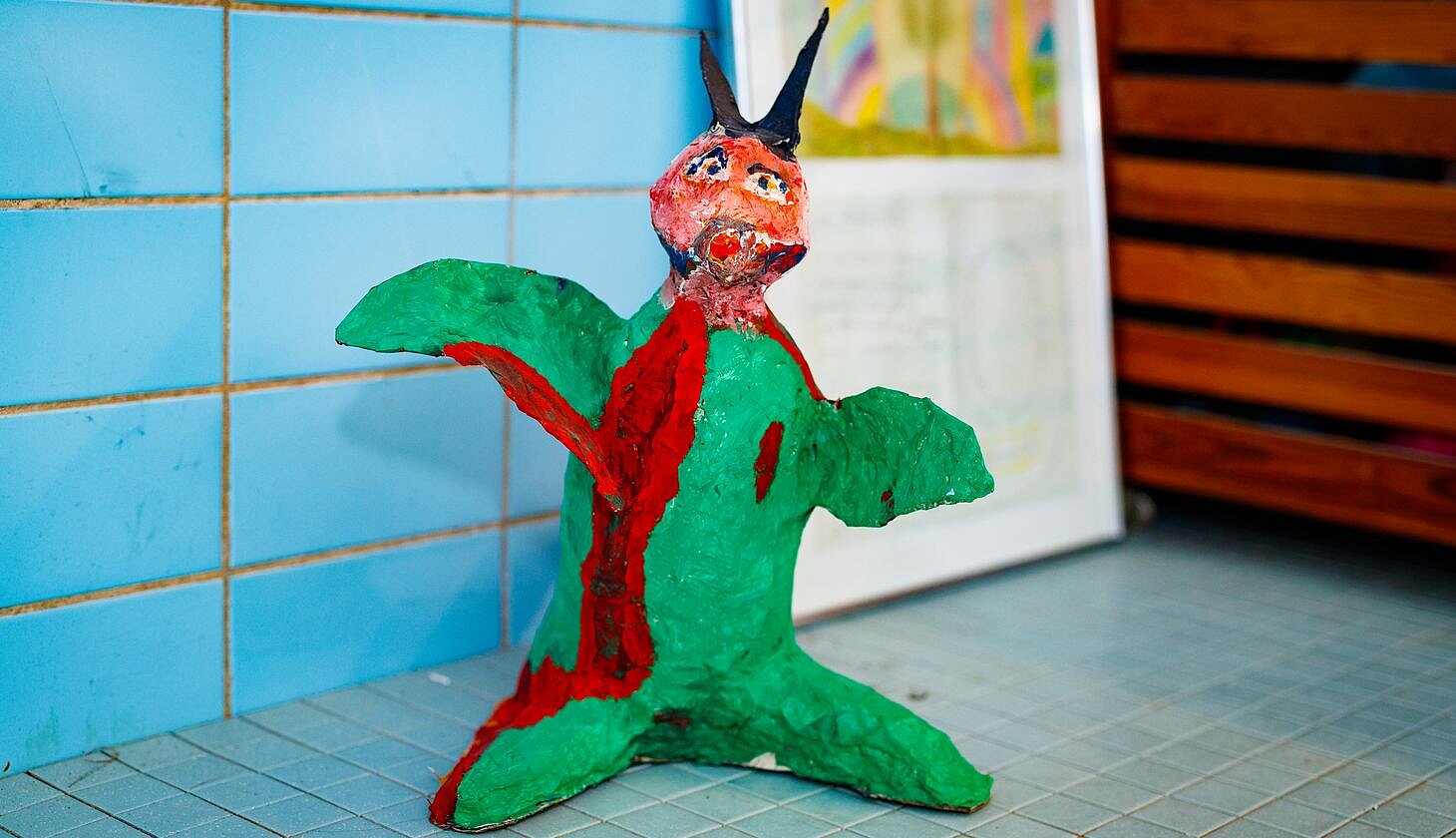 Eine farbenfrohe, handgefertigte Skulptur steht auf einem gefliesten Boden vor einer blau gefliesten Wand mit Kunstwerken im Hintergrund.