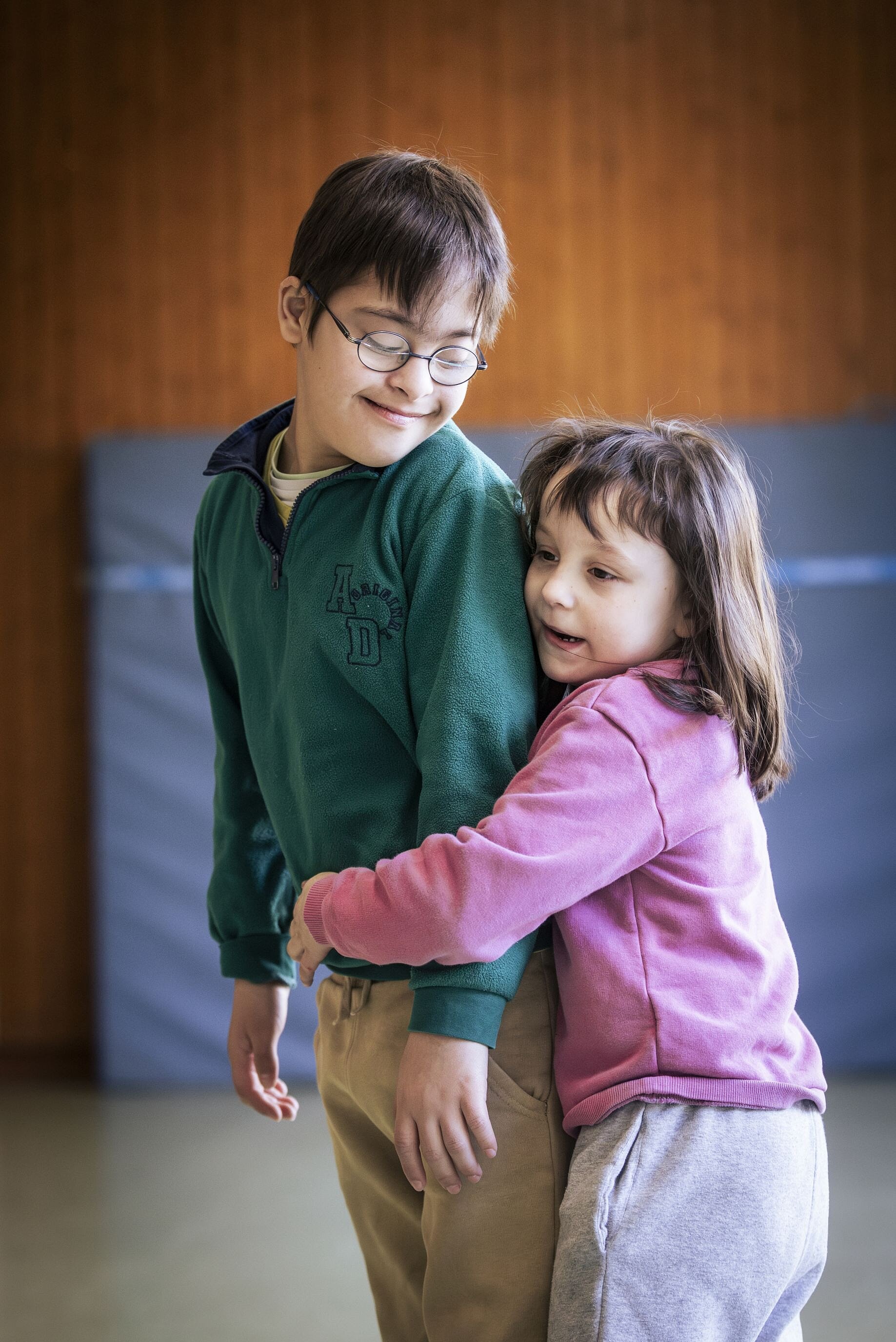 Ein Mädchen umarmt einen Jungen liebevoll von der Seite. Beide Kinder lächeln und stehen eng beieinander.