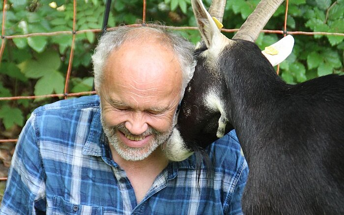Ein lächelnder Mann der auf dem Boden sitzt und mit einer Ziege kuschelt