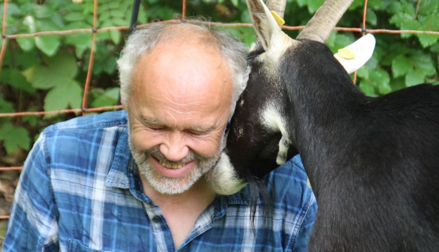 Ein lächelnder Mann der auf dem Boden sitzt und mit einer Ziege kuschelt
