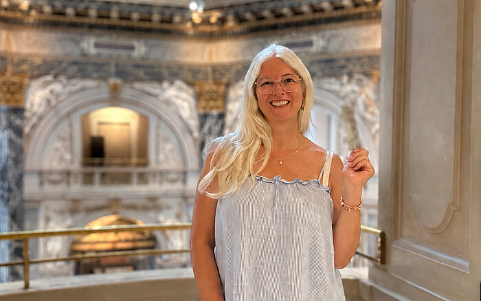 Eine lächelnde Frau mit langen blonden Haaren und Brille steht auf einer Empore vor der prunkvollen Innenarchitektur eines historischen Gebäudes in Wien.