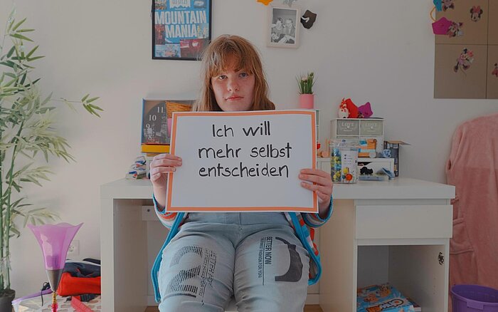 Eine junge Person mit mittellangen Haaren sitzt in einem bunt eingerichteten Zimmer und hält ein Schild mit der Aufschrift „Ich will mehr selbst entscheiden“.
