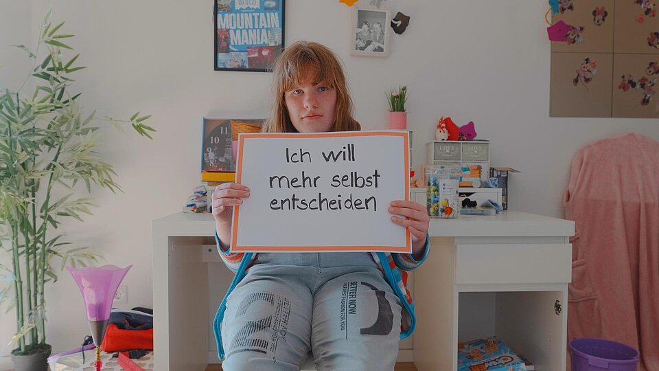 Eine junge Person mit mittellangen Haaren sitzt in einem bunt eingerichteten Zimmer und hält ein Schild mit der Aufschrift „Ich will mehr selbst entscheiden“.