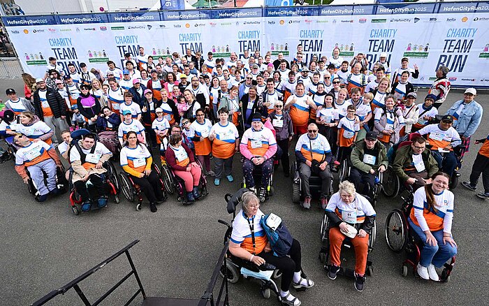 Große Gruppe von Menschen – einige im Rollstuhl, andere stehend – posieren beim HafenCity Run 2025 für ein Gruppenfoto. Alle tragen Sozialkontor-Shirts in Weiß, Orange und Blau.