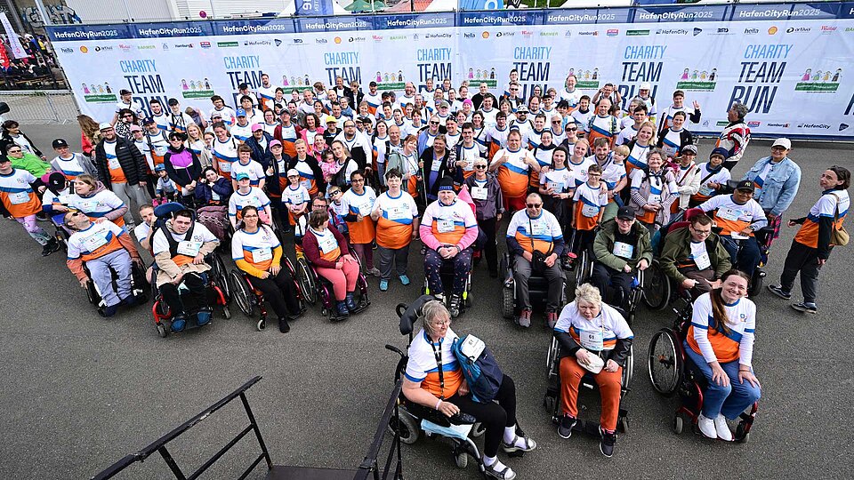 Große Gruppe von Menschen – einige im Rollstuhl, andere stehend – posieren beim HafenCity Run 2025 für ein Gruppenfoto. Alle tragen Sozialkontor-Shirts in Weiß, Orange und Blau.
