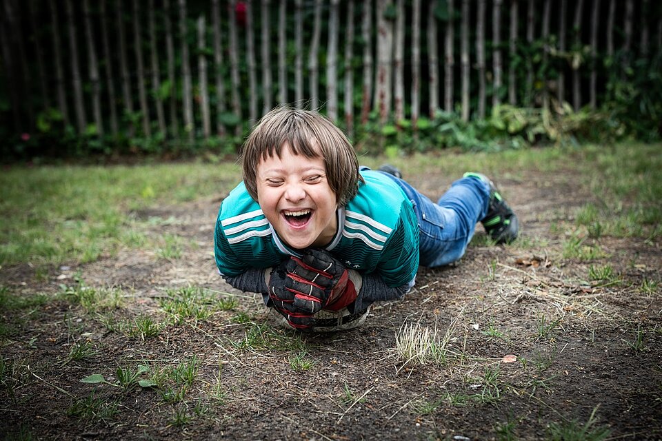 Ein junge liegt lachend auf einen Fußball am Boden auf einem Sportplatz.