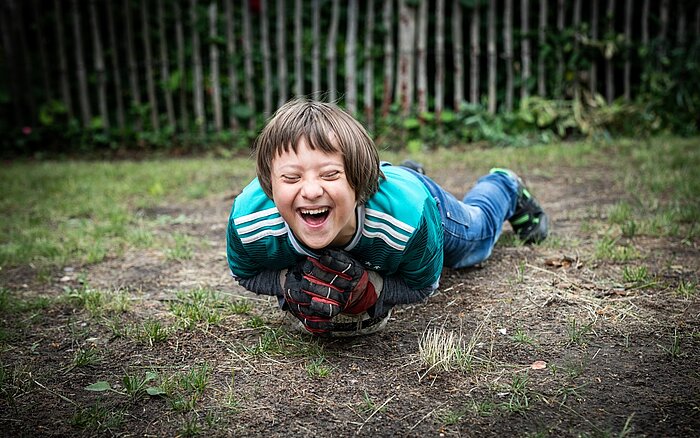 Ein junge liegt lachend auf einen Fußball am Boden auf einem Sportplatz.