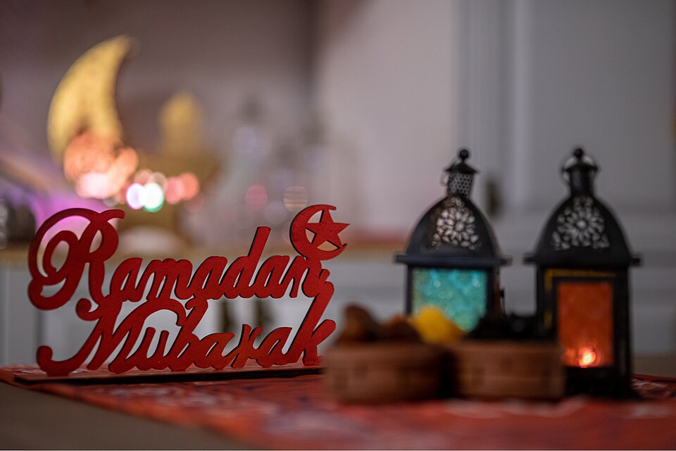 Dekoratives Arrangement mit zwei orientalischen Laternen und einem Schriftzug aus Holz, der „Ramadan Mubarak“ zeigt. Im Hintergrund leuchten unscharfe Lichter in warmen Farben.